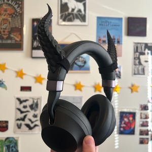 May include: Black over-ear headphones with decorative black horns attached to the headband. The headphones have a matte finish and are held up against a wall decorated with posters and star-shaped lights.