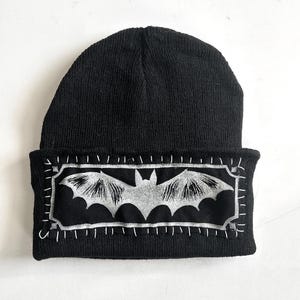 May include: A black knit beanie featuring a rectangular patch sewn onto the cuff with white thread. The patch is black with a silver-colored graphic of a bat with spread wings, framed by a silver border and decorative corner accents.