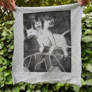May include: A gray, square textile featuring a black and white illustration of a three-headed dog standing on a rocky surface. The artwork has a linocut style, with the dog's form and the background rendered in solid black.