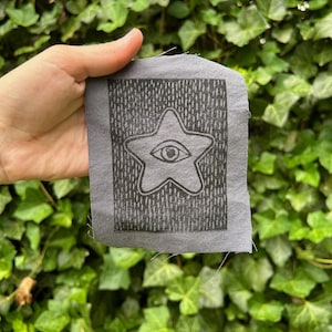 May include: A gray fabric patch featuring a star with an eye design. The star is outlined in black, with a detailed eye in the center. The background is a textured pattern. The patch is held against a backdrop of green foliage.