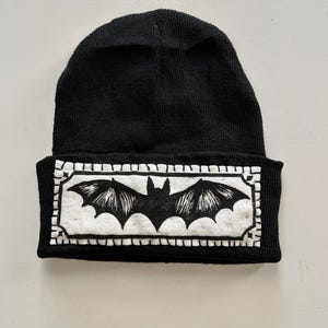 May include: Black knitted beanie hat with a white rectangular patch displaying a black bat illustration. The bat has outstretched wings and is centered on the patch. The patch has a decorative border.