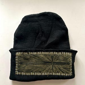 May include: Black knit beanie with a rectangular patch featuring a sword design in olive green. The patch is stitched onto the beanie with white thread. The beanie is a winter accessory.