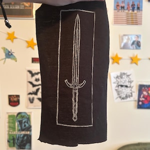 May include: A black fabric item featuring a white, hand-drawn sword design within a rectangular border. The sword has a detailed hilt and blade. The item is held up against a wall with posters and star-shaped lights.