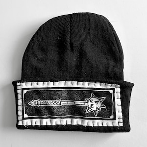 May include: Black beanie hat with a white rectangular patch featuring a mace design. The mace has a spiked ball head and a detailed handle. The patch is bordered with a white stitched design.