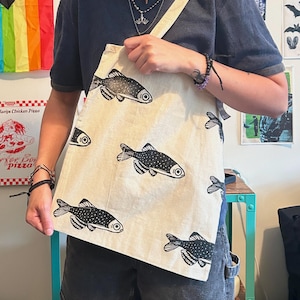 May include: A beige canvas tote bag featuring a repeating black ink print of fish. The fish illustrations are oriented in various directions across the fabric surface. The bag includes a single shoulder strap and a rectangular body construction.