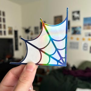 May include: A holographic sticker in the shape of a spider web corner. The sticker has a white background with a web design outlined in dark blue. The edges of the sticker have a rainbow sheen.