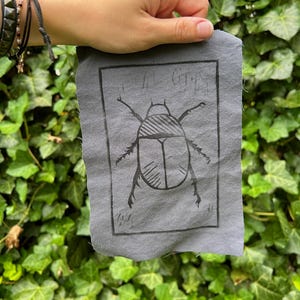 May include: A gray fabric square featuring a black illustration of a beetle within a rectangular frame. The beetle has detailed legs and antennae. The fabric has a raw, unfinished edge.