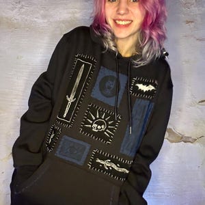 May include: Black hoodie with multiple embroidered patches. Patches include a sword, skull, eye, and bat. The patches are stitched onto the hoodie with white thread. The hoodie has a front pocket and a drawstring hood.