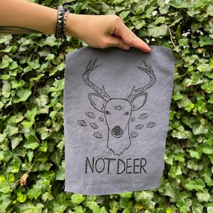 May include: A gray rectangular fabric with a black illustration of a deer head with multiple eyes and antlers. The text "NOT DEER" is printed below the deer illustration. The fabric is held in front of a green leafy background.