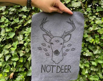 May include: A gray rectangular fabric with a black illustration of a deer head with multiple eyes and antlers. The text "NOT DEER" is printed below the deer illustration. The fabric is held in front of a green leafy background.