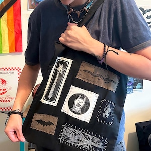 May include: A black tote bag featuring several rectangular and circular fabric patches sewn onto the front. The patches display white and brown screen-printed graphics including a mace, a sword, a bat, a spider, a stylized animal head, and a cross.