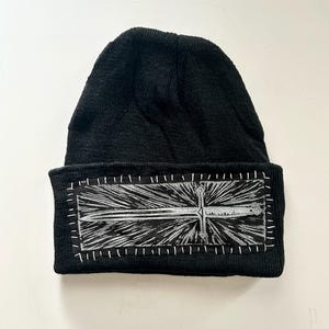 May include: Black knit beanie with a rectangular patch featuring a silver sword design. The patch is stitched onto the beanie with white thread, creating a contrasting border. The sword design has a detailed hilt and blade.