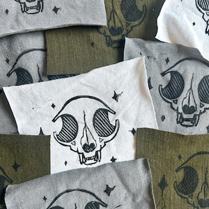 May include: A collection of rectangular fabric patches in shades of gray, olive green, and off-white. Each patch features a black ink screen-printed graphic of a stylized animal skull surrounded by small four-pointed star shapes.