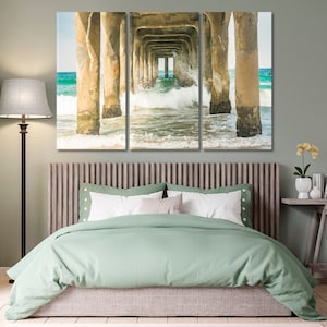May include: A three-panel canvas print of a pier over the ocean. The image shows the underside of the pier with waves crashing through the supports. The water is a turquoise blue and the sky is a light blue. The pier is made of concrete and the supports are painted white.