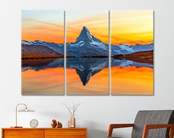 May include: A panoramic view of a mountain peak reflected in a still lake at sunset. The sky is a vibrant orange and yellow, with clouds reflecting in the water. The mountain is snow-capped and appears to be in the Swiss Alps.