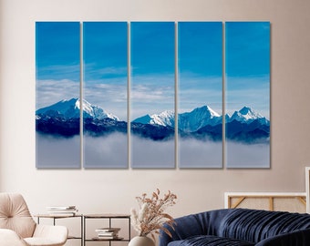 May include: A panoramic view of snow-capped mountains with a layer of fog in the foreground. The sky is a bright blue with white clouds.