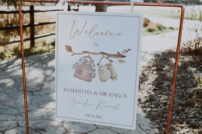 May include: A white sign with brown text that reads "Welcome to Samantha & Michael's Gender Reveal May 14, 2022". The sign features a watercolor illustration of a branch with two baby clothes hanging from it.