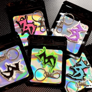 May include: Keychains in black pouches with holographic inserts. Each keychain features a unique, stylized design in various colors, including pink, purple, green, and blue. The keychains have silver-colored clasps. The text "NOCTURNAL PRINT SHOP" is visible.