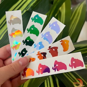 May include: A set of six sheets of holographic stickers featuring a silhouette of a mammoth in various colors, including green, blue, orange, and pink. The stickers are arranged in a fan shape and are held in a hand. The stickers are labeled "Nocturnal Print Shop".