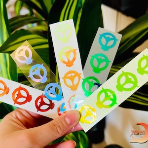 May include: Five sheets of holographic stickers with a repeating design of a stylized alien head. The stickers are in various colors, including green, blue, orange, yellow, and pink.