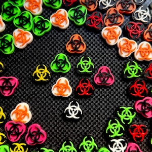 May include: A collection of colorful, biohazard symbol charms in various colors including orange, green, pink, red, yellow, and white. The charms are arranged on a textured black surface. These charms are ideal for crafting and jewelry making.