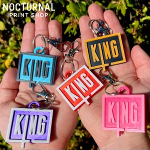 May include: Five rectangular keychains featuring the word KING in raised lettering. The keychains come in blue, orange, yellow, purple, and pink, each attached to a silver metal lobster clasp. The design includes a vertical line extending from the letter G.