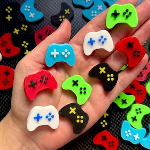 May include: A collection of colorful miniature game controllers in various colors, including blue, red, green, black, and white. Each controller features a cross-shaped directional pad and button details. The controllers are scattered on a dark surface.