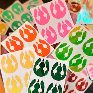 May include: A collection of colorful stickers featuring a stylized design. The stickers are in various colors, including pink, orange, green, and yellow. The design is a white outline of a shape with three rounded elements. The stickers are on a white background.