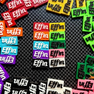 May include: A collection of small, rectangular objects in various colors, each bearing the word "Effin" in a contrasting color. The objects are arranged on a dark, textured surface, with a few grouped together and others scattered around.