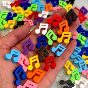 May include: A hand holding a variety of colorful plastic music note charms. The charms are in various colors, including red, blue, green, yellow, orange, purple, black, and white.