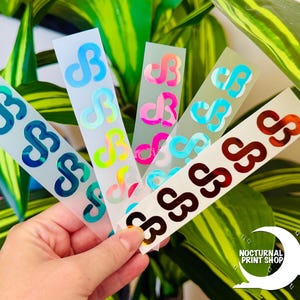 May include: A hand holds several sticker strips with a repeating abstract design in various colors, including blue, pink, and brown. The stickers have a holographic effect. The logo for Nocturnal Print Shop is in the bottom right corner.