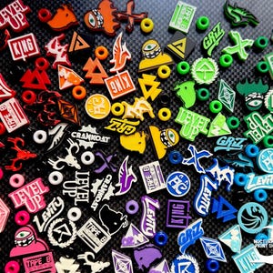 May include: A collection of colorful shoe charms with various designs and text, including "LEVEL UP," "KING," and "LEVITY." The charms are in shades of red, orange, yellow, green, blue, and purple, with white accents. The background is a textured black surface.