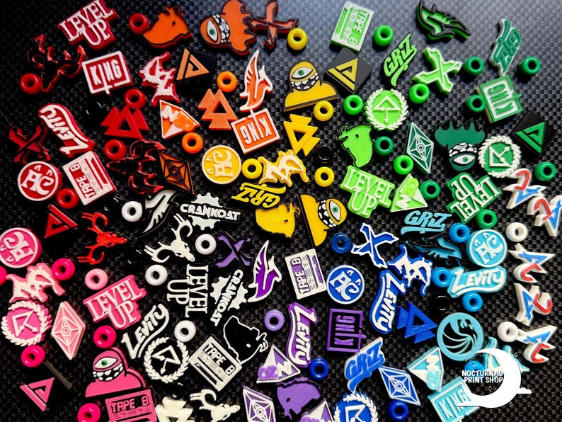 May include: A collection of colorful shoe charms with various designs and text, including "LEVEL UP," "KING," and "LEVITY." The charms are in shades of red, orange, yellow, green, blue, and purple, with white accents. The background is a textured black surface.