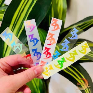 May include: A hand holding several colorful holographic stickers. Each sticker features Japanese characters in various iridescent colors, including blue, red, orange, and green. The stickers are rectangular and have a transparent background.