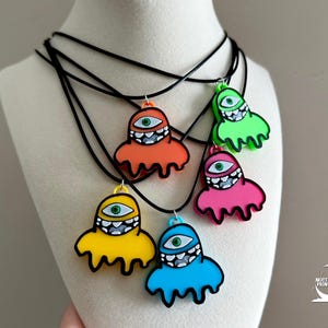 May include: A collection of colorful monster-shaped pendant necklaces. Each pendant is a different color: orange, green, pink, yellow, and blue. The monsters have one eye and a jagged mouth. The necklaces have black cords.