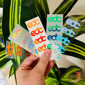 May include: Five sheets of holographic stickers with the letters "edc" repeated on each sheet. The sheets are in different colors: blue, green, orange, pink, and rainbow.