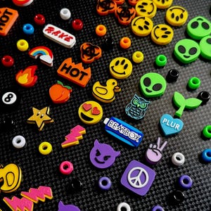 May include: A collection of colorful plastic charms and small round beads featuring various designs including yellow stars, orange peaches, green alien faces, yellow smiley faces, blue hearts with the text PLUR, and blue rectangles with the text BEATBOX.