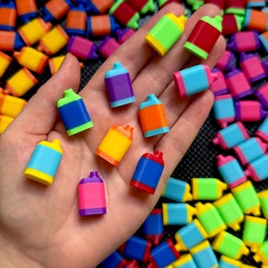 May include: A hand holding several colorful miniature spray paint bottles. The bottles are various colors, including blue, green, yellow, orange, red, and purple. The background is filled with more of the same miniature bottles.