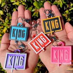 May include: Five colorful keychains with the word "KING" printed on them. Each keychain is a different color: blue, yellow, red, pink, and purple. The keychains are held in a person's hand.