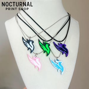 May include: Five necklaces featuring stylized bird-shaped pendants in white, green, purple, pink, and light blue. Each pendant hangs from a thin black or silver chain. The text NOCTURNAL PRINT SHOP is visible in the upper left corner of the image.