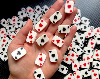 May include: A collection of white playing card charms with red hearts, black clubs, and spades. The charms are rectangular and have a glossy finish. Some charms are held in a hand, while others are scattered around.