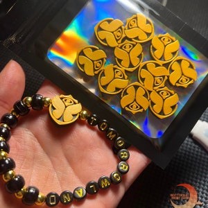 May include: A black beaded bracelet with gold lettering spelling "TOMORROW" and a gold charm with a stylized eye design. The bracelet is in a hand, and a bag of gold charms with the same eye design is in the background.