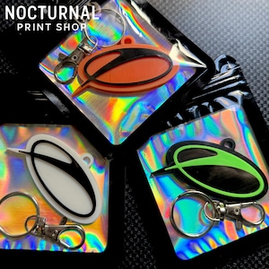 May include: Three keychains in individual holographic pouches, each featuring an oval-shaped emblem with a stylized black letter A. The emblems are orange, white, and neon green. Each keychain includes a silver metal split ring and a lobster clasp.
