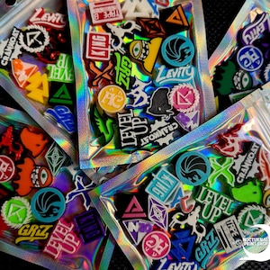 May include: Assortment of colorful, graphic shoe charms in clear, holographic zip bags. Charms feature various shapes, logos, and text like "LEVEL UP", "LEVITY", and "CRANKOAT". The charms are in various colors including red, blue, green, and yellow.