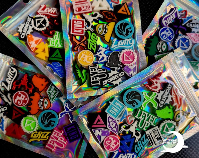 May include: Assortment of colorful, graphic shoe charms in clear, holographic zip bags. Charms feature various shapes, logos, and text like "LEVEL UP", "LEVITY", and "CRANKOAT". The charms are in various colors including red, blue, green, and yellow.