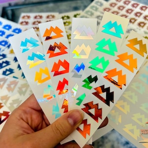 May include: Assortment of iridescent sticker sheets featuring a repeating pattern of geometric triangles in various colors, including orange, blue, green, and silver.
