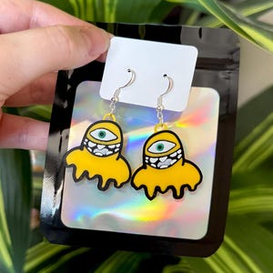 May include: A pair of dangle earrings featuring a yellow, blob-like creature with a single green eye and a wide mouth filled with white teeth. The earrings are attached to a white display card inside a black, holographic-finish plastic pouch.