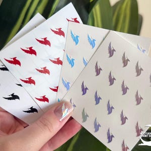 May include: A hand holds several sheets of stickers with repeating bird designs. The stickers feature birds in red, black, blue, and iridescent colors. The background includes green leaves and the logo for Nocturnal Print Shop.