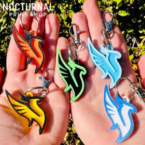 May include: Five phoenix-shaped keychains with metal lobster claw clasps and split rings. The keychains feature vibrant color combinations: orange and black, lime green and black, light blue, yellow and black, and blue and white. Text reads NOCTURNAL PRINT SHOP.