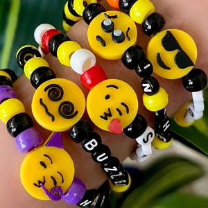 May include: Five colorful bracelets with yellow smiley face charms and black and white letter beads. The bracelets spell out "BUZZ" and "HASH".